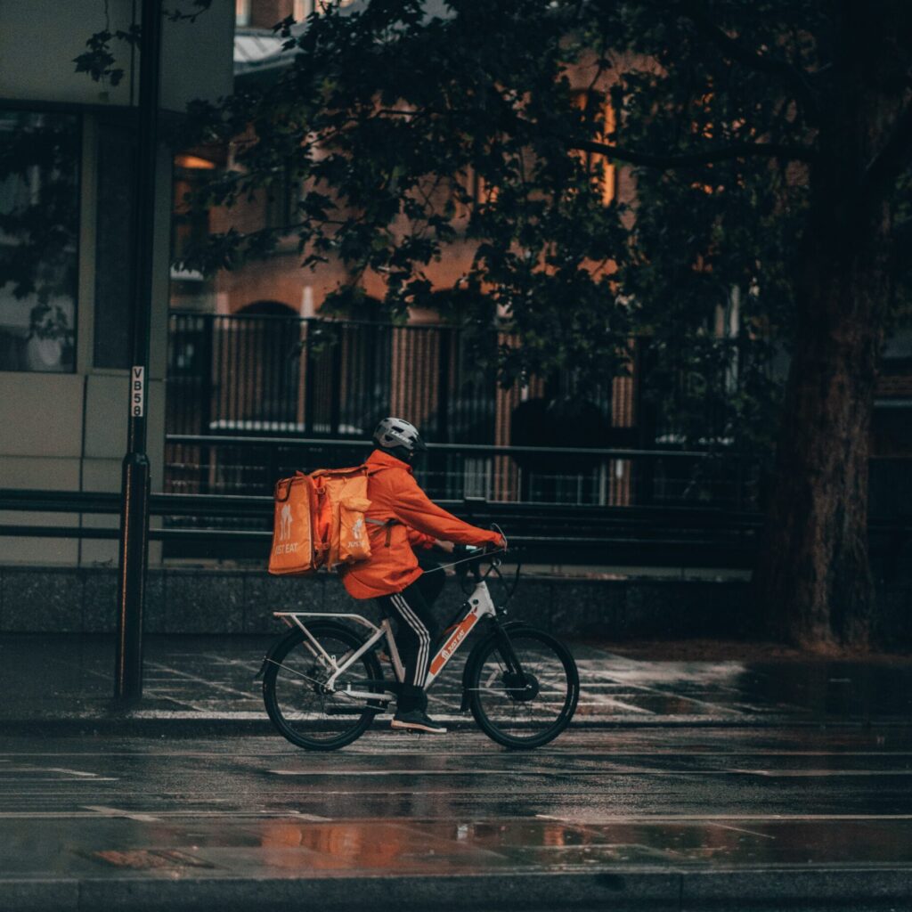 Foto: Joshua Lawrence (unsplash.com)

Mann mit Orangener Jacke und orangenem großen Lieferrucksack fährt auf nasser Fahrbahn über eine städtische Straße.